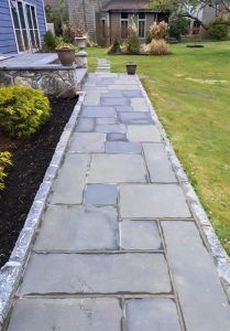 A stone-paved walkway leads to a house porch, bordered by grass and a garden bed with a small shrub.