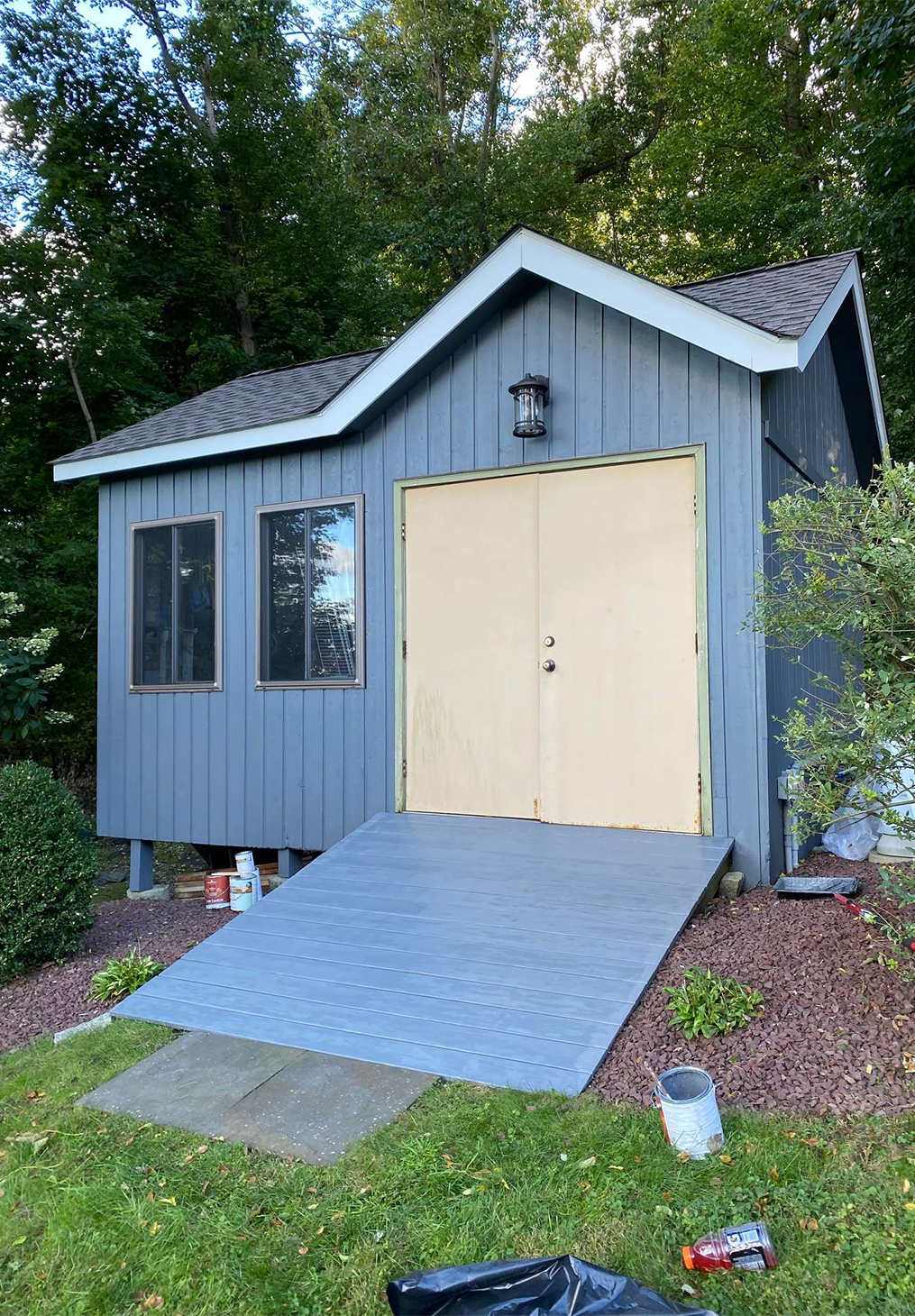 A gray shed with double doors and a blue ramp, set in a yard with green trees and landscaping gravel.