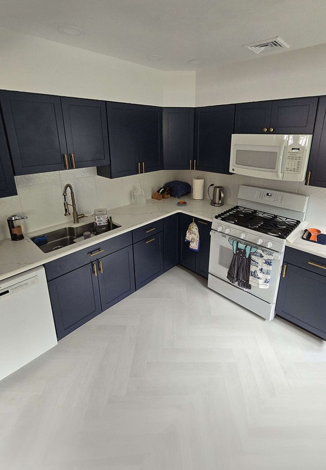 A modern kitchen with navy blue cabinets, white countertops, stainless steel sink, and white herringbone floor tiles.