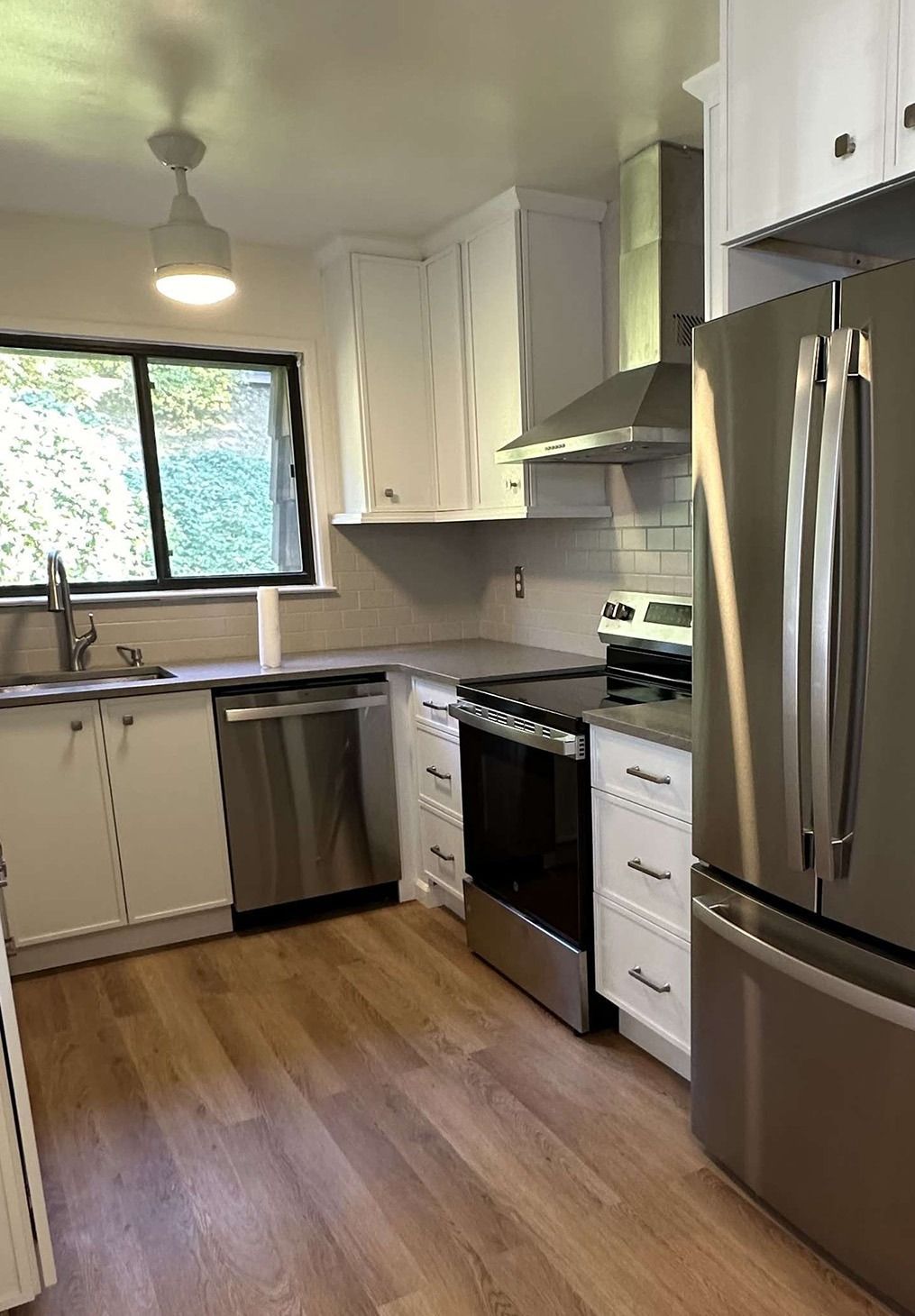 A modern kitchen with white cabinets, stainless steel appliances, light wood floors, and a window above the sink.