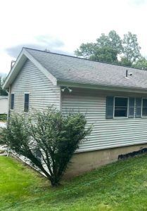 A light-colored, single-story house with horizontal siding and a gray roof, surrounded by green grass and a bush.