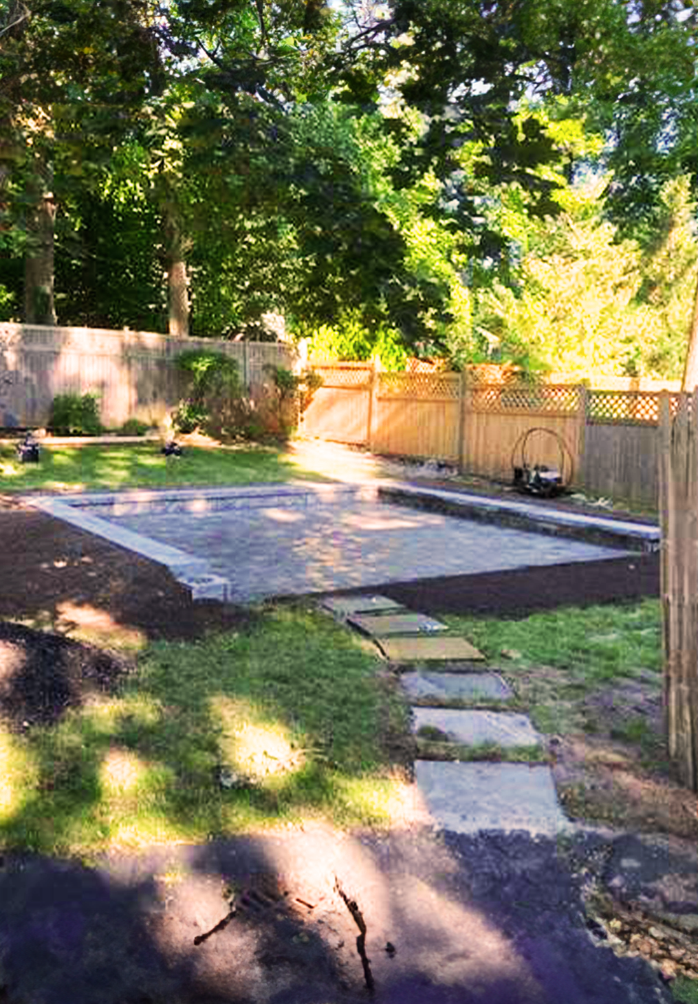 A rectangular stone patio under construction in a grassy backyard, featuring a stepping stone path and wooden fence.