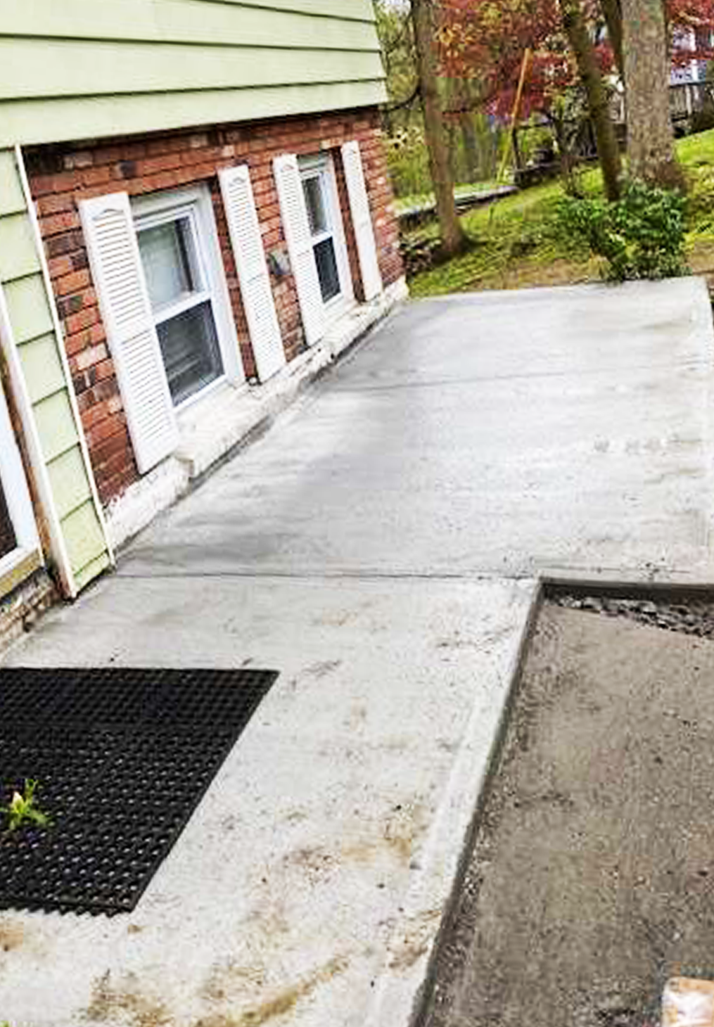 A freshly poured concrete patio adjacent to a light green house with brick accents and white-shuttered windows.