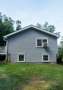 Light blue, side-gabled house with three windows and an air conditioning unit, situated in a green, grassy yard.