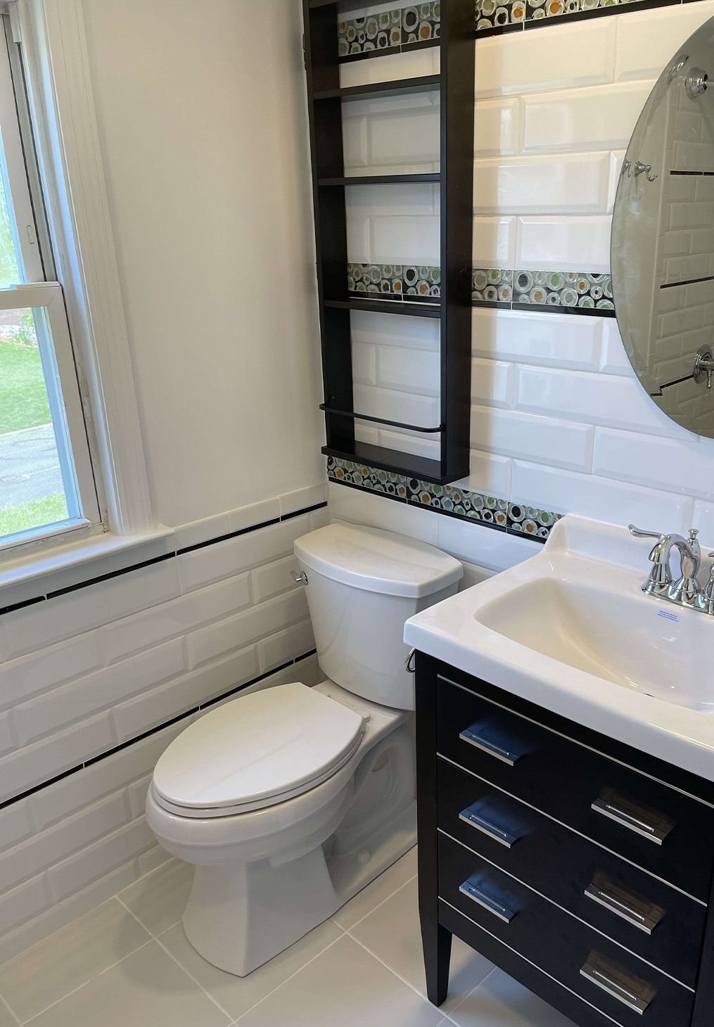 A small bathroom with a white toilet, black vanity, white subway tile walls with decorative trim, and a wall shelf.