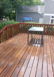 A wooden deck with a square metal table, overlooking a backyard with a blue umbrella and a grey house.