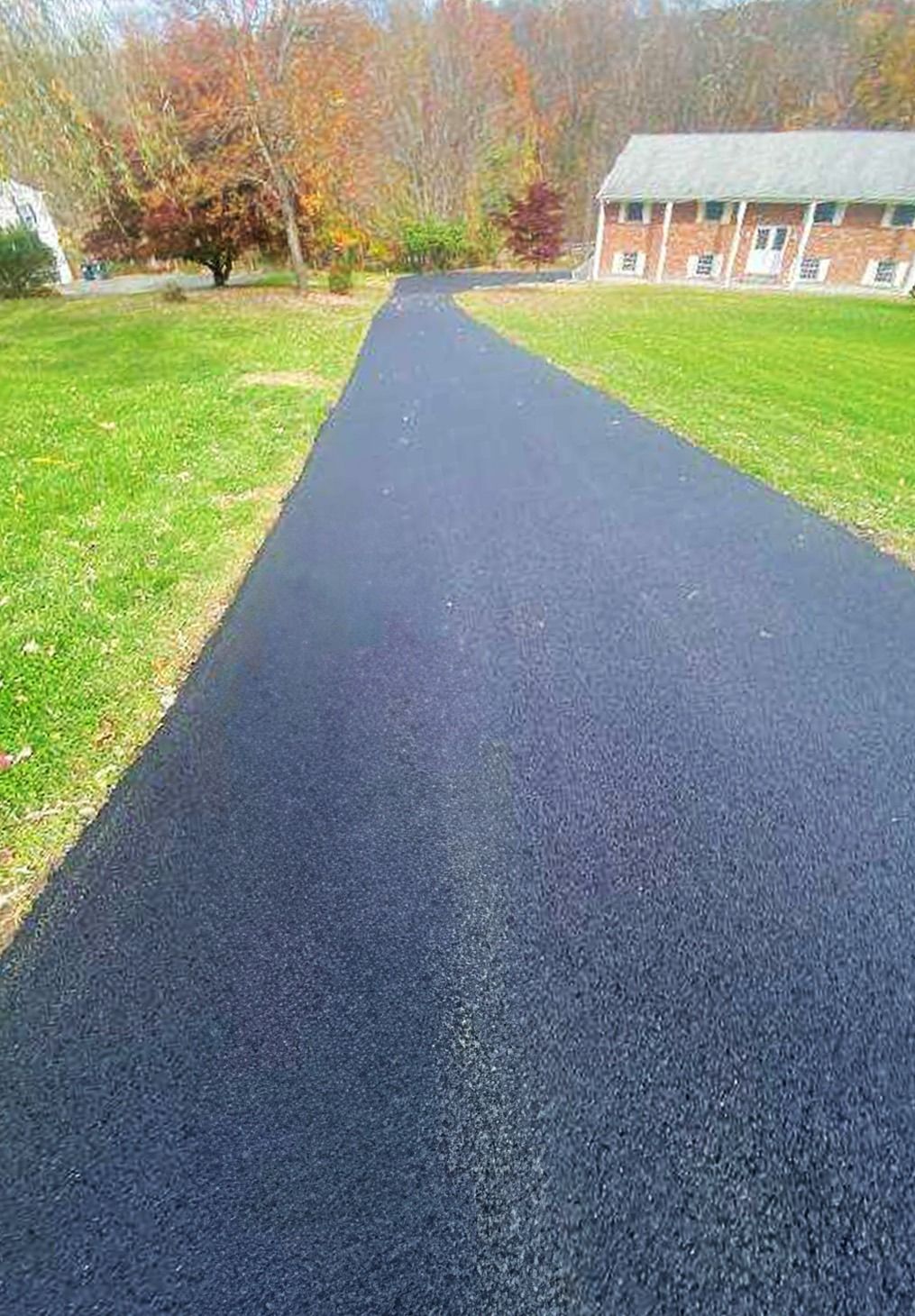 A freshly paved, dark asphalt driveway leads toward a brick house surrounded by trees with autumn foliage.