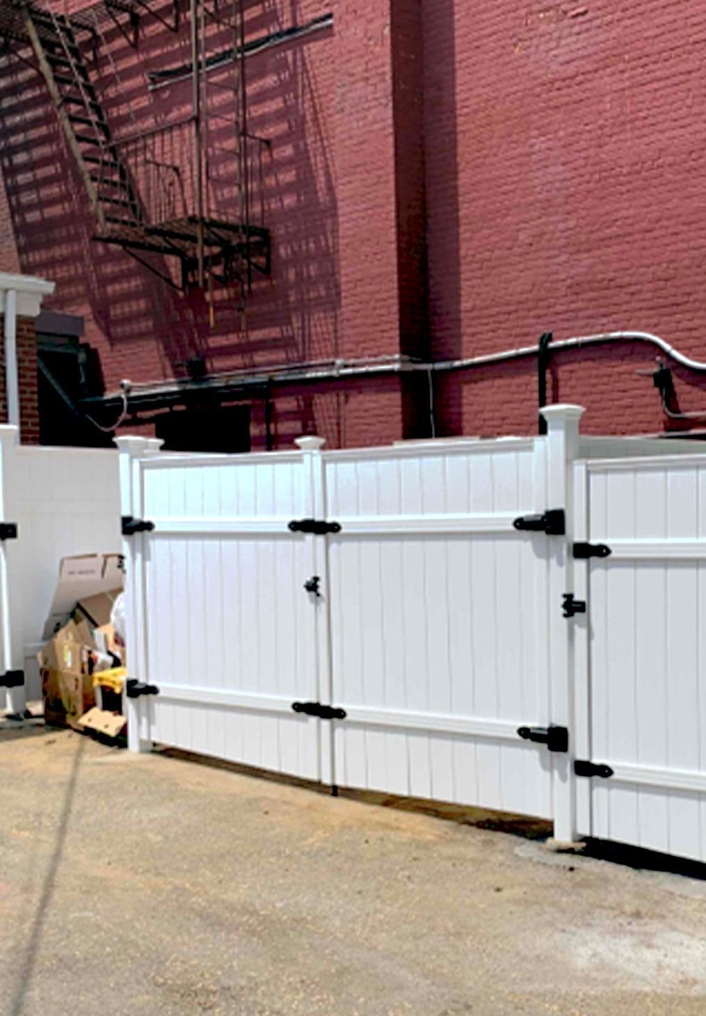 A white vinyl fence with black hardware stands in front of a red brick building with a metal fire escape.