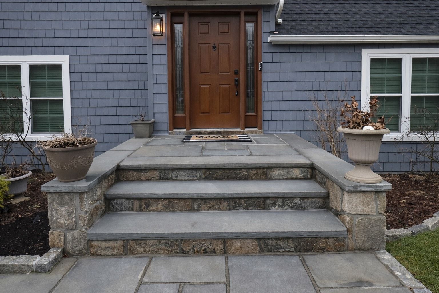 A blue-sided home with a wooden front door, slate-colored stone steps, and two planters on a front stone patio.