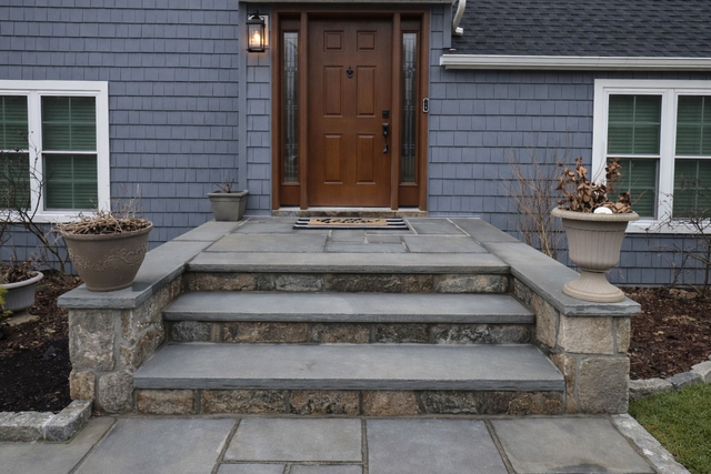 A set of three gray stone steps leading up to a wooden front door, flanked by stone walls with potted plants.