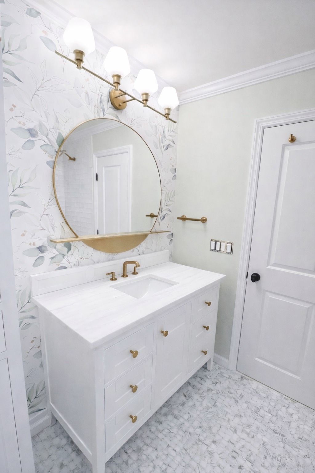 A white bathroom vanity with gold hardware, a round gold mirror, and floral wallpaper against a soft green wall.