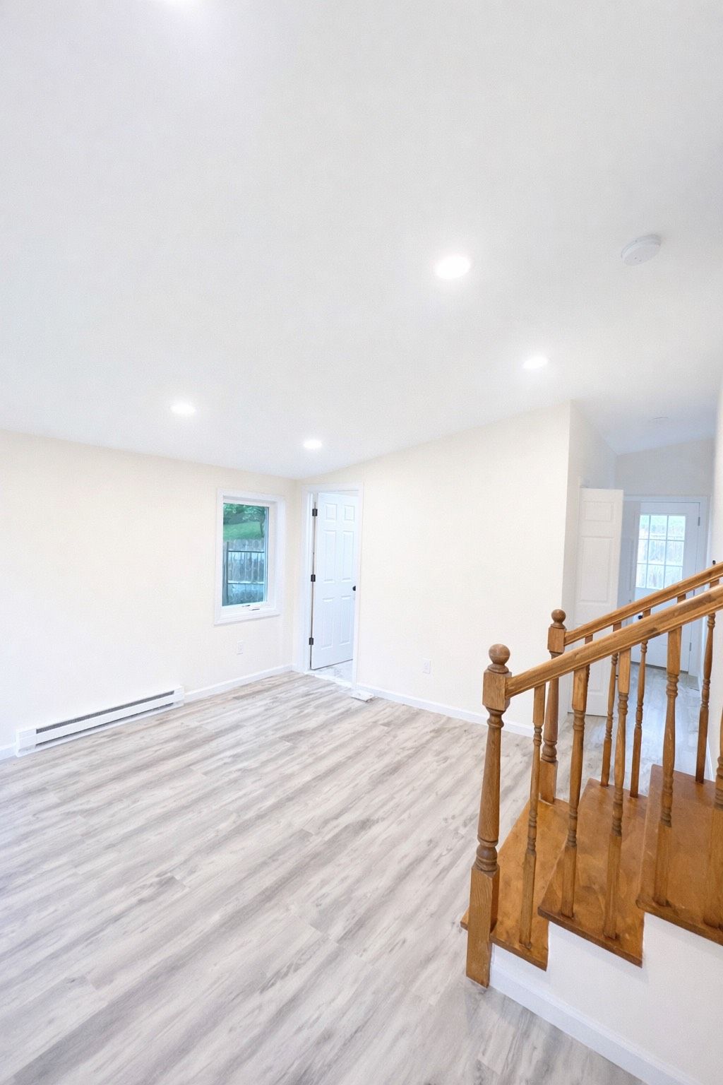 A bright, modern interior room with light wood-patterned flooring, white walls, recessed lighting, and wooden stairs.