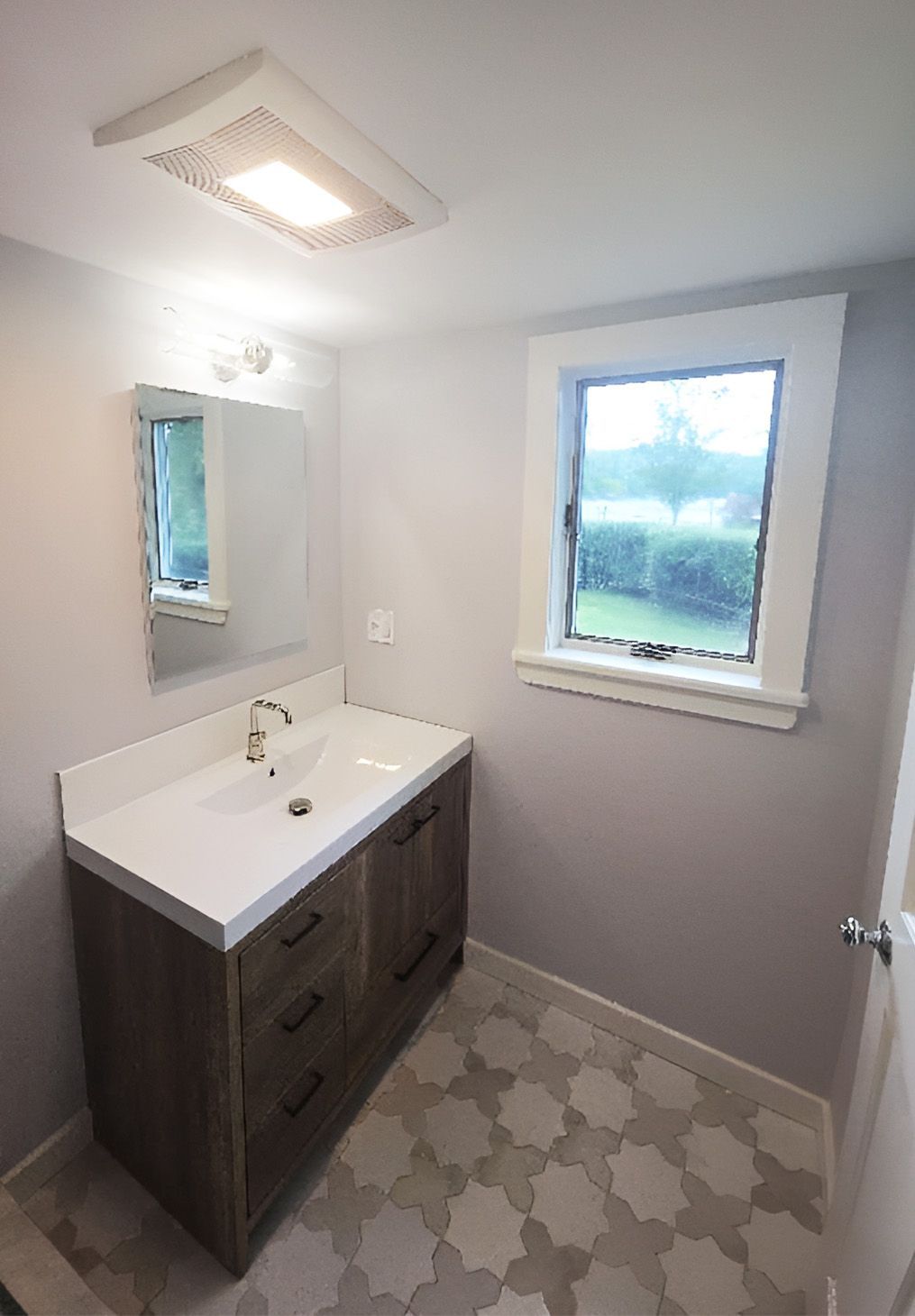 A modern bathroom featuring a white sink vanity with a wood cabinet, a wall mirror, a window, and patterned tile flooring.
