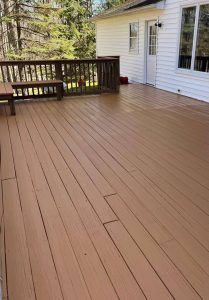 A clean, brown wooden deck attached to a white house with a railing, a bench, and a window, viewed on a sunny day.