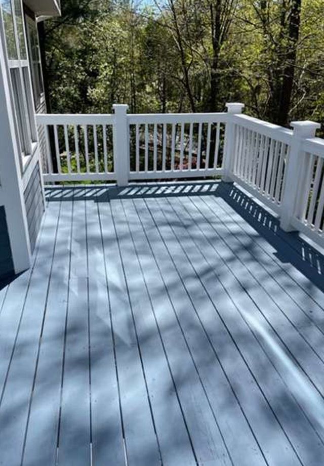 A light blue wooden deck with white railings, featuring shadows from surrounding trees on a sunny day.