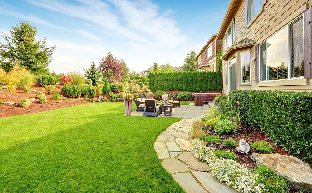 Landscaped backyard with green lawn, stone path, patio seating, and a house with hedges and flower beds