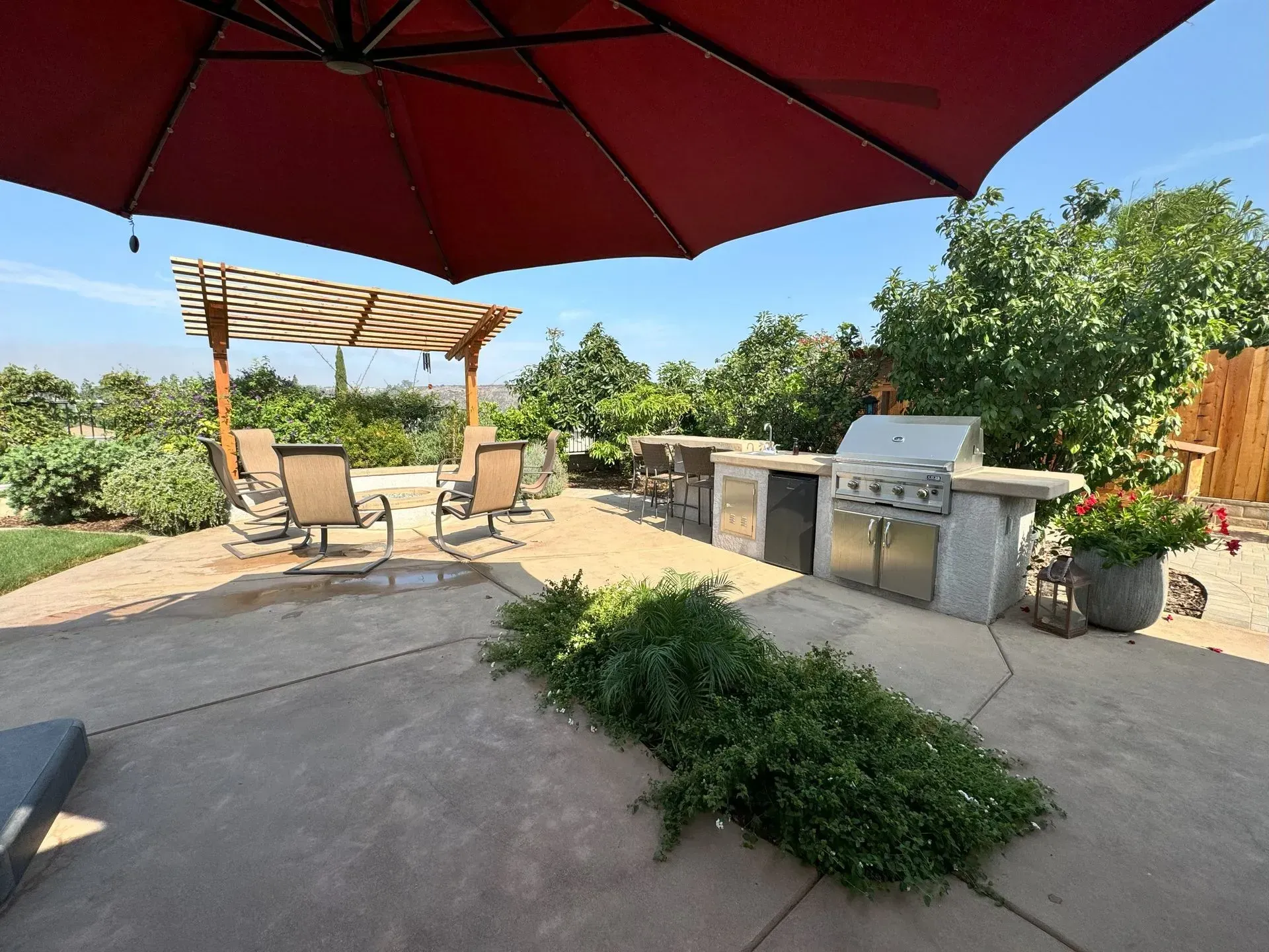 Patio with outdoor kitchen, seating area, pergola, and red umbrella on a sunny day.
