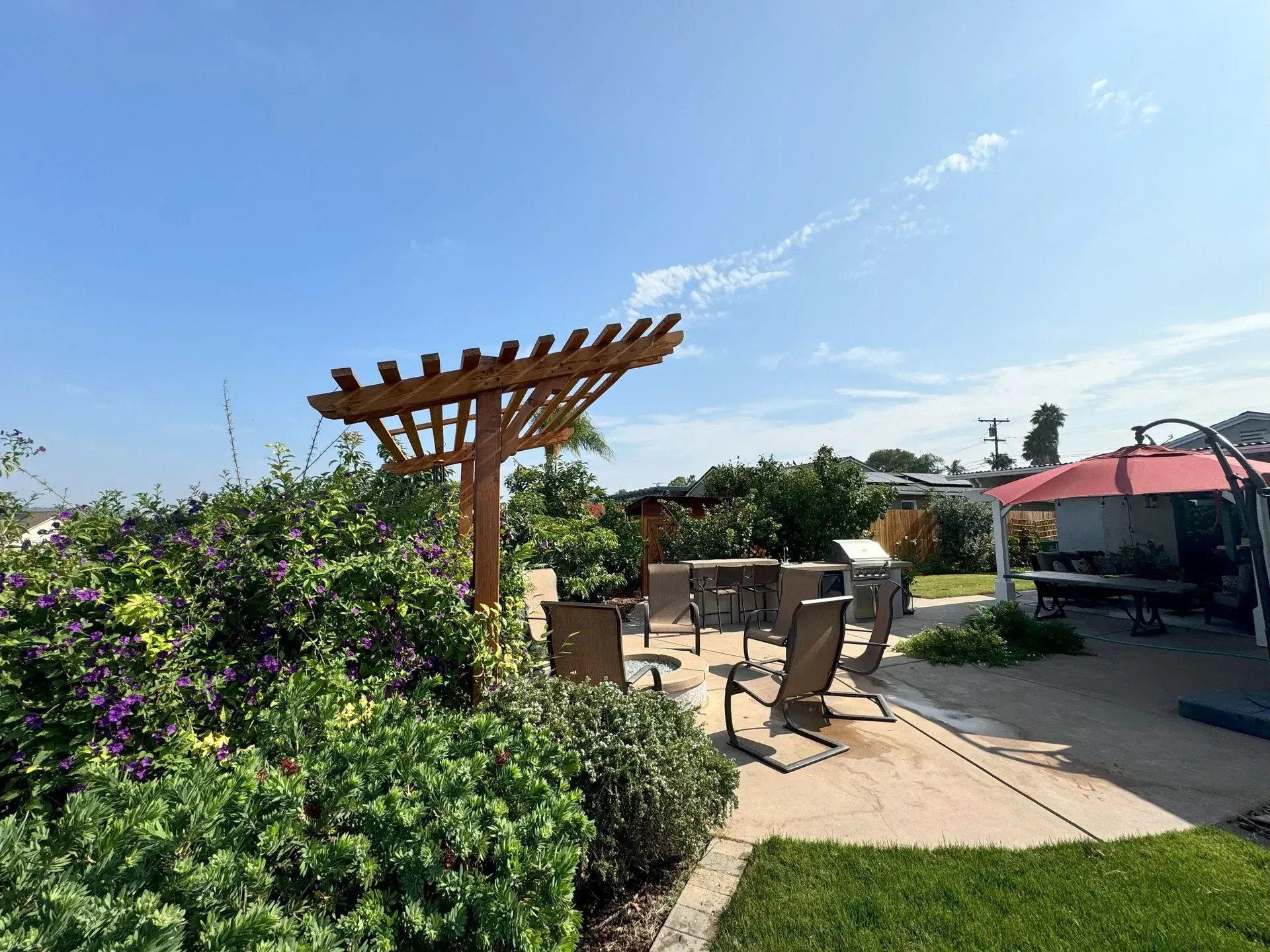 Patio with wooden pergola, chairs, tables, and green bushes under a blue sky.