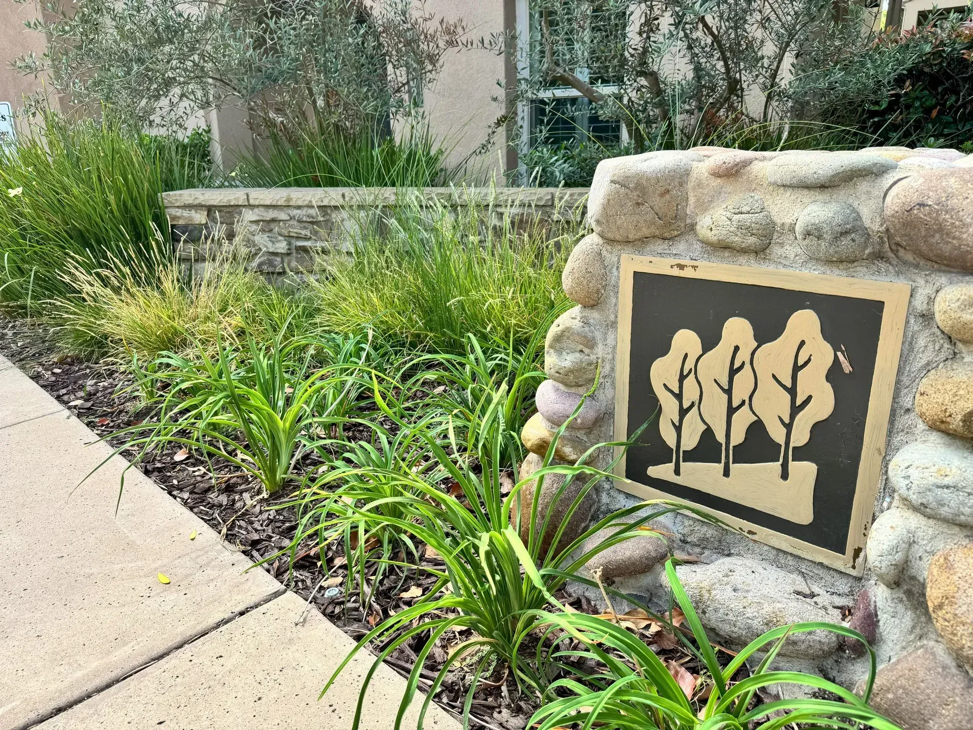 Sign with tree silhouettes in a landscaped area next to a sidewalk.