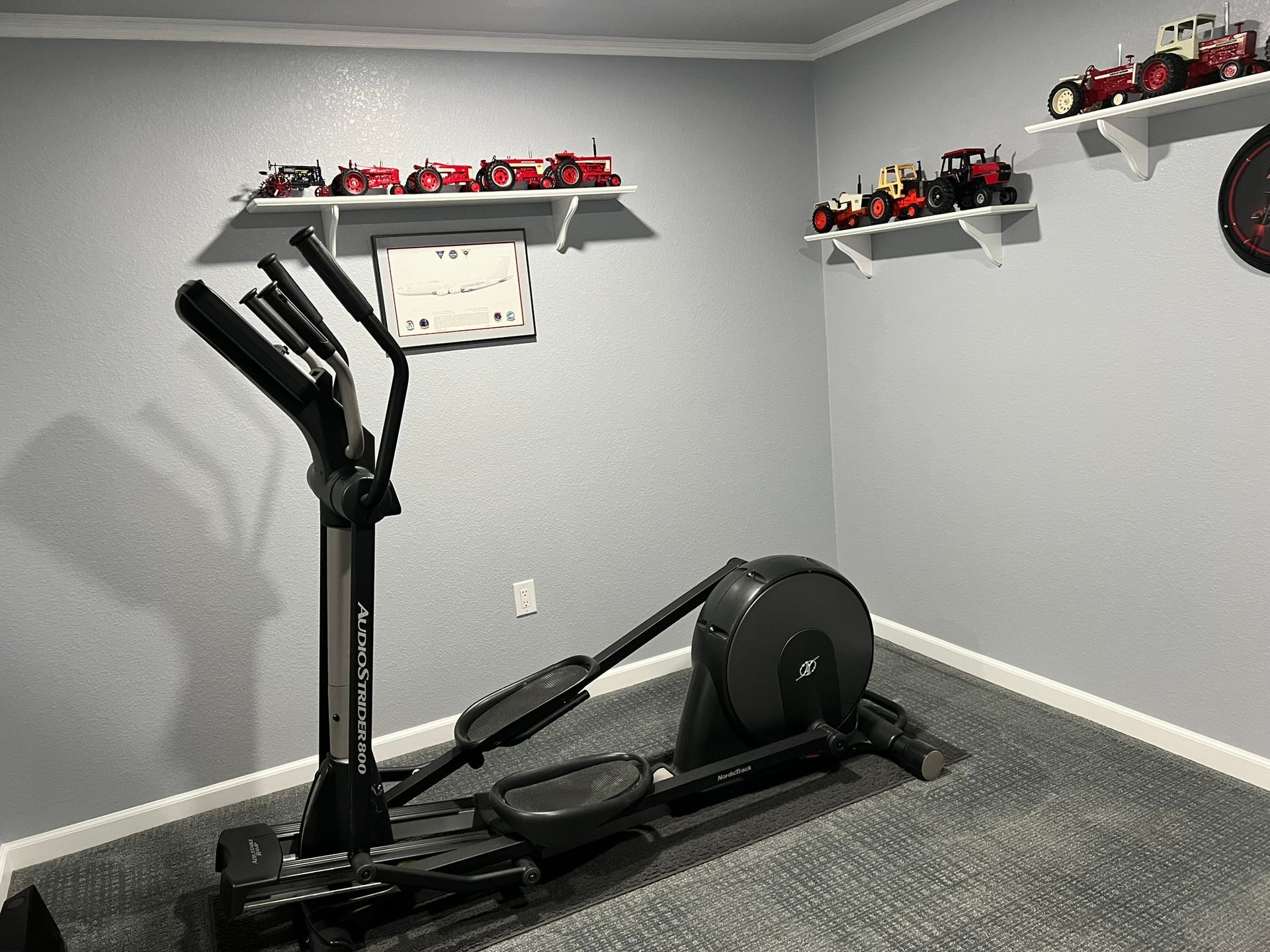 An elliptical machine is sitting in a room next to a shelf with tractors on it.