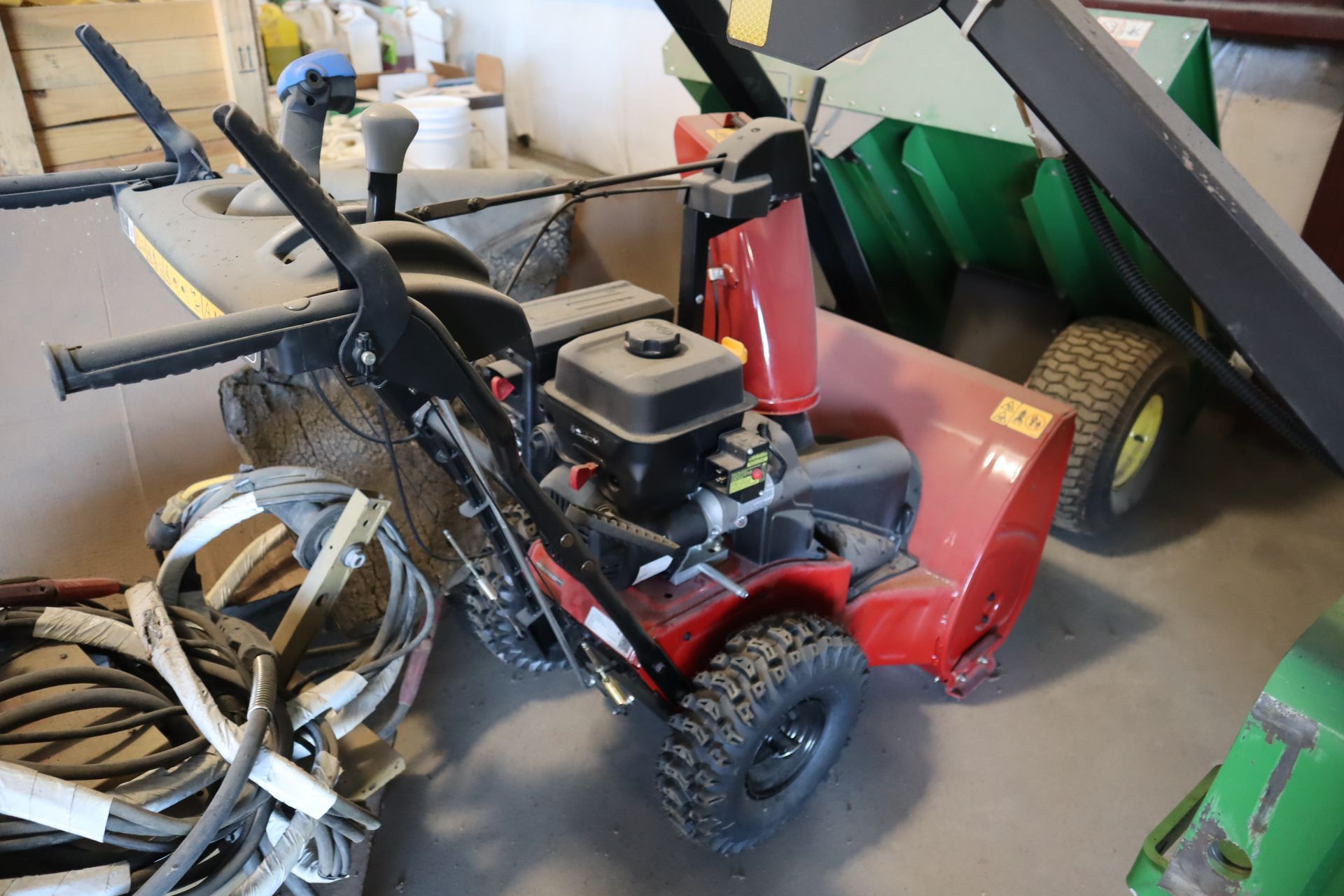 A red snow blower is sitting in a garage next to a green tractor.