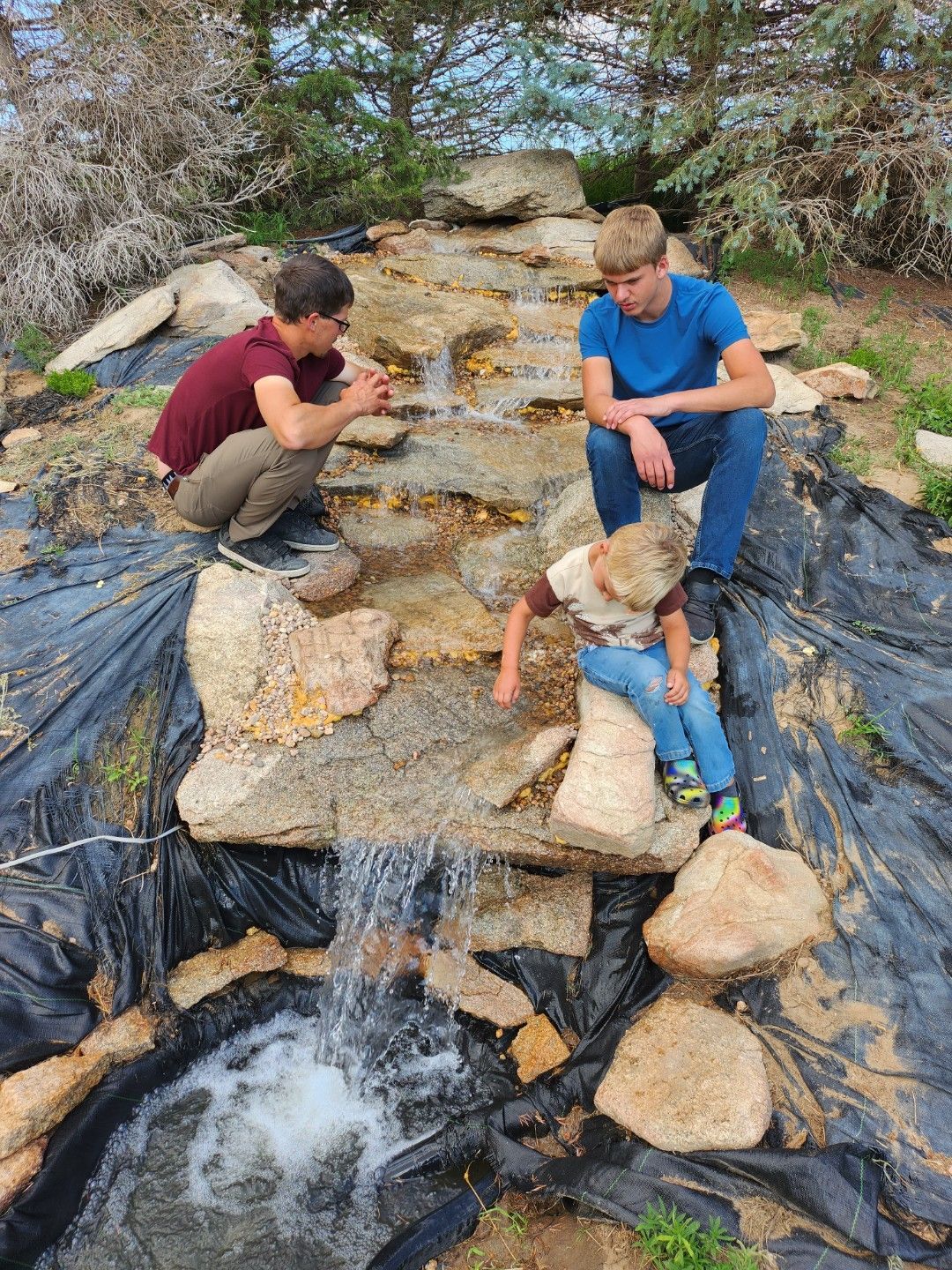Two boys are sitting on rocks next to a waterfall.
