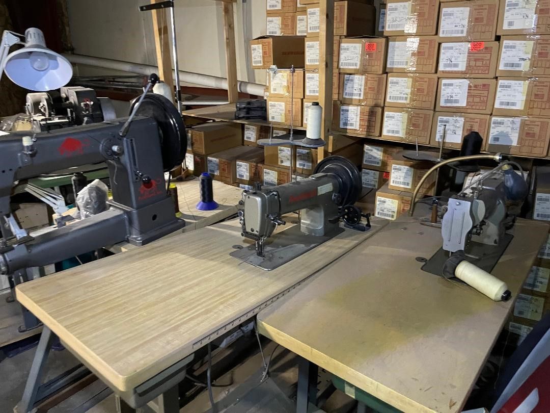 Three industrial sewing machines on tables in a workshop, with boxes in the background.