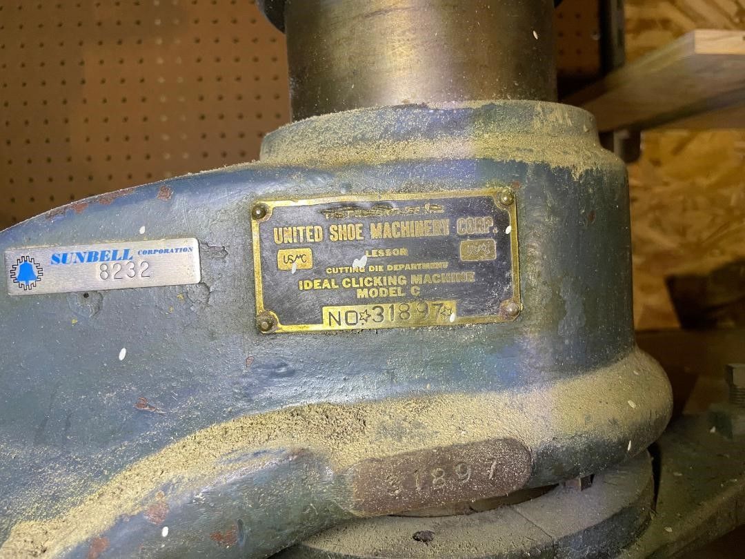 Close-up of a dusty blue drill press, with a brass manufacturer's plate that says 