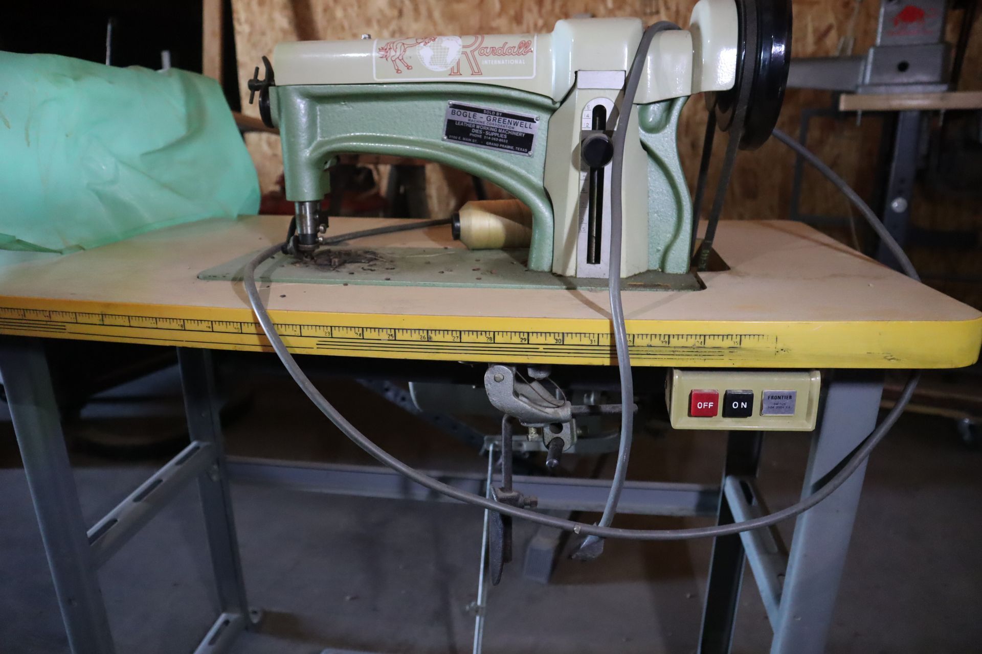 A heavy-duty sewing machine on a yellow table. The machine has a light green and beige color scheme, set in a workshop.