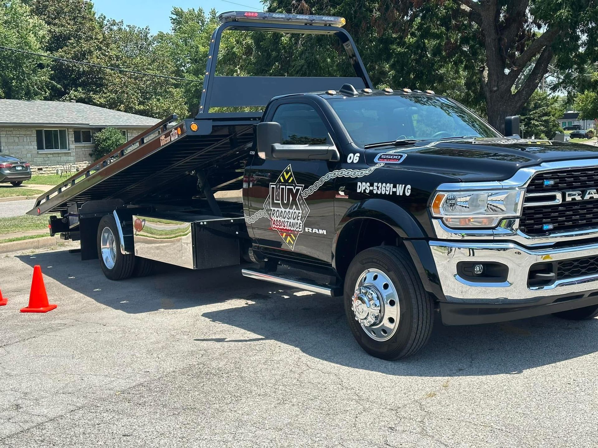 A black flatbed tow truck parked on an asphalt lot in front of a house, with orange safety cones in the foreground.