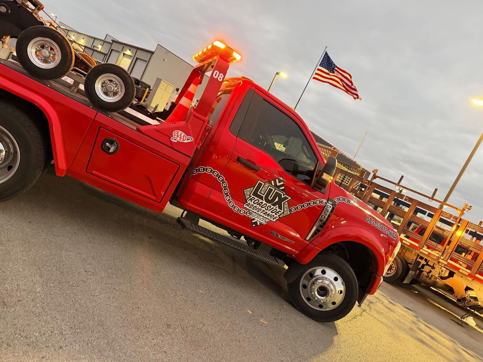 A bright red tow truck with a graphic on its door parked in a lot under a cloudy sky with an American flag visible.