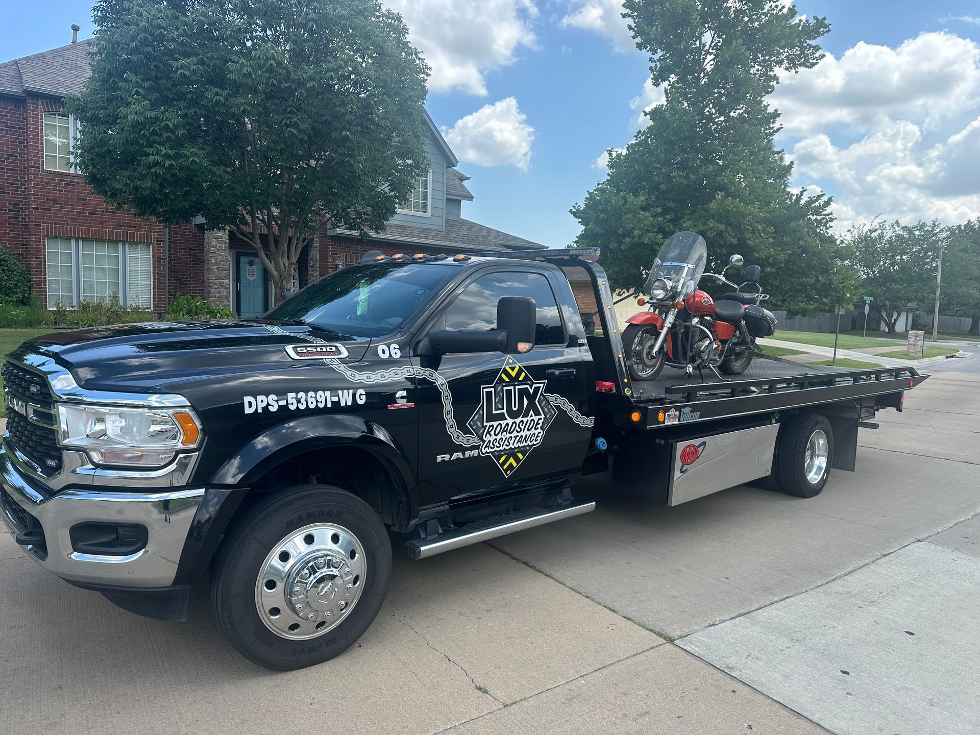 A black flatbed tow truck with a motorcycle loaded on the back, parked on a residential street in front of a brick house.