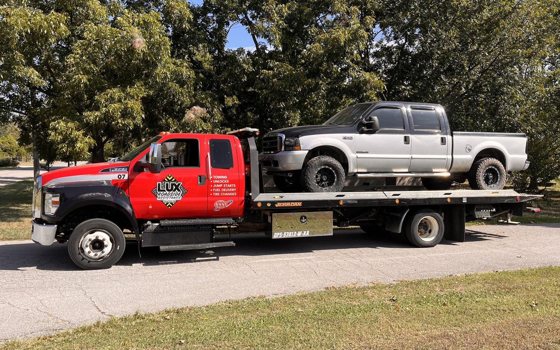 A red flatbed tow truck carries a silver pickup truck along a road lined with trees.