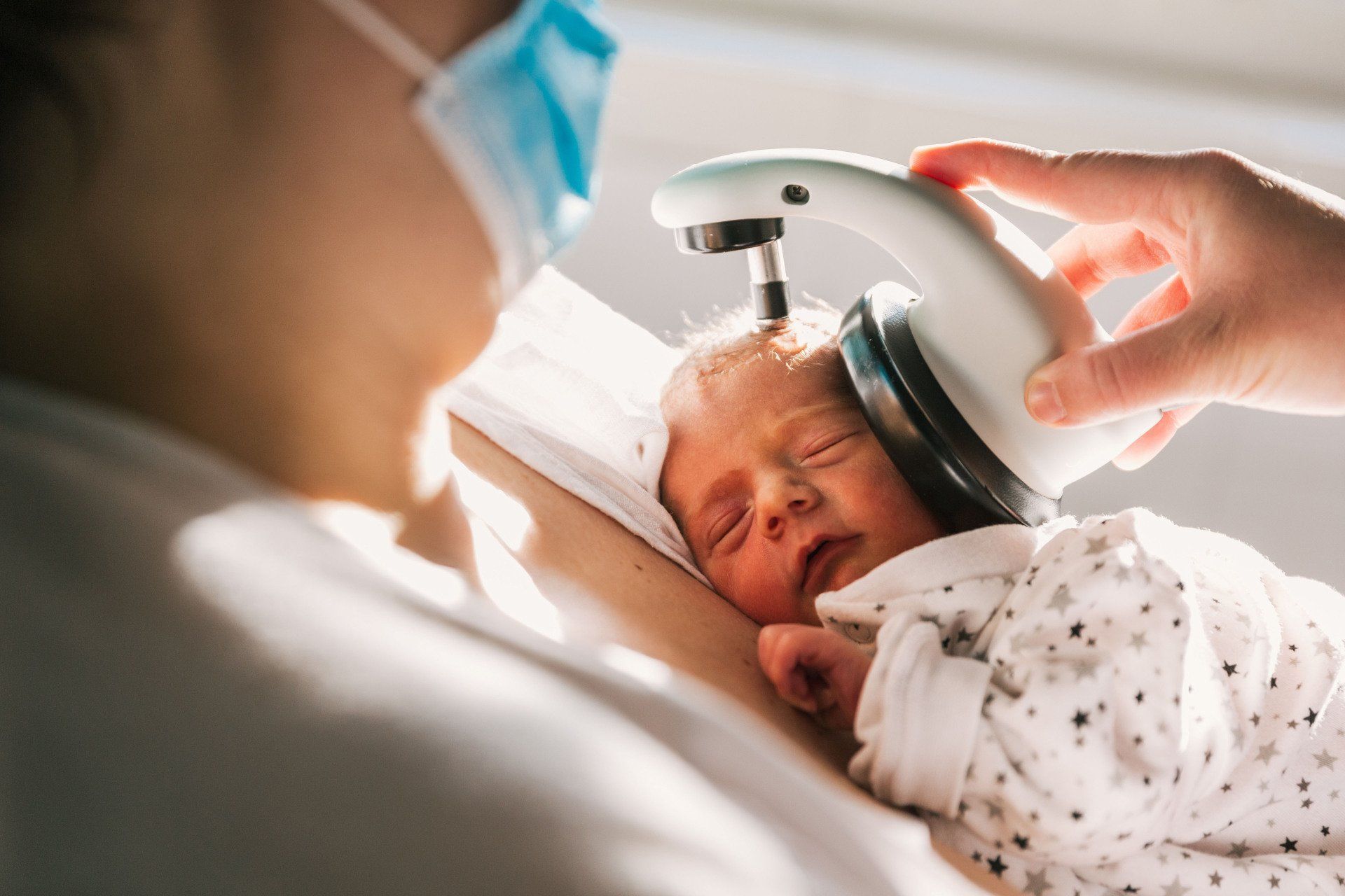 A medical professional uses a handheld device to scan the head of a newborn resting against a person wearing a mask.