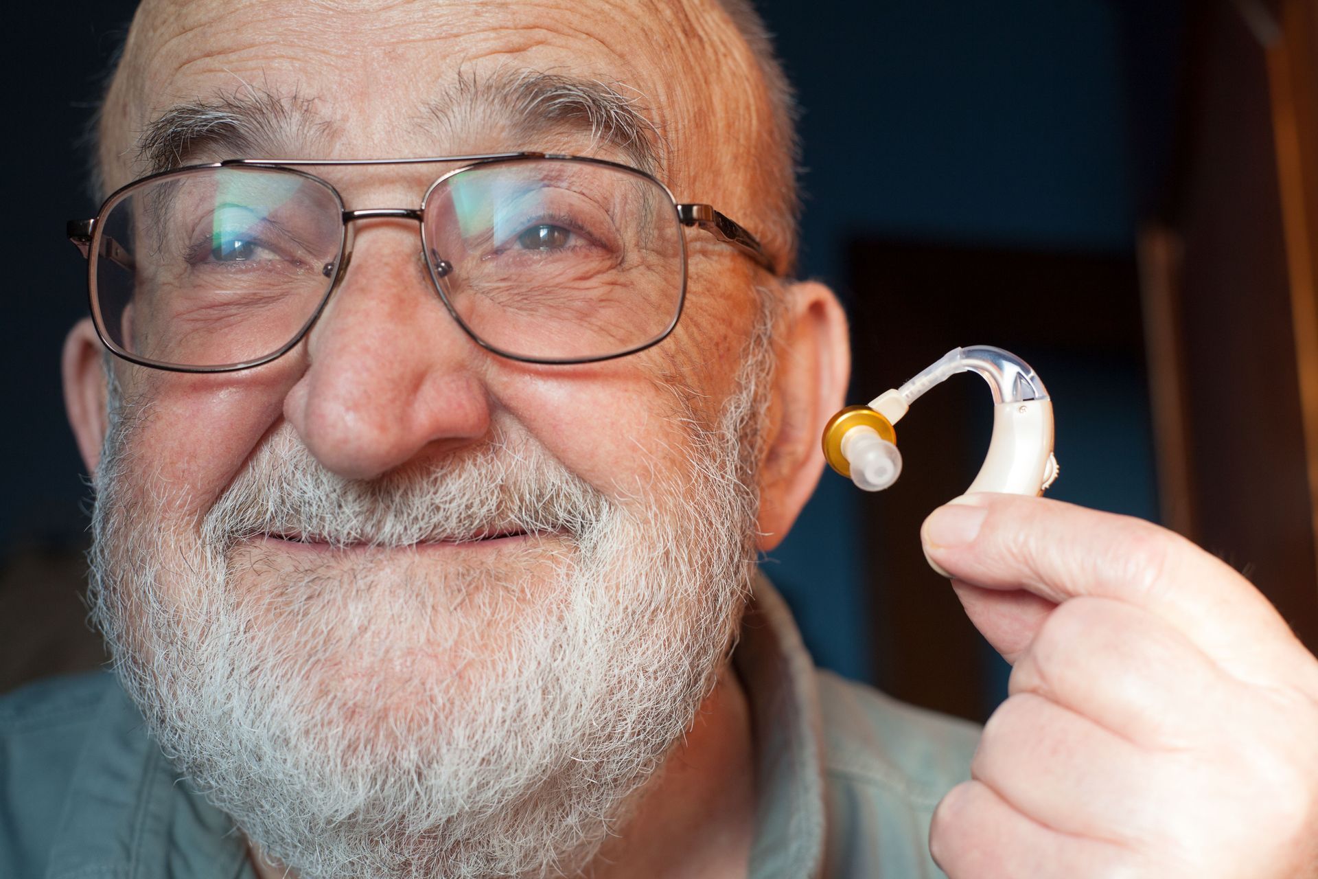 A smiling person holds a small, beige behind-the-ear hearing aid near their ear.