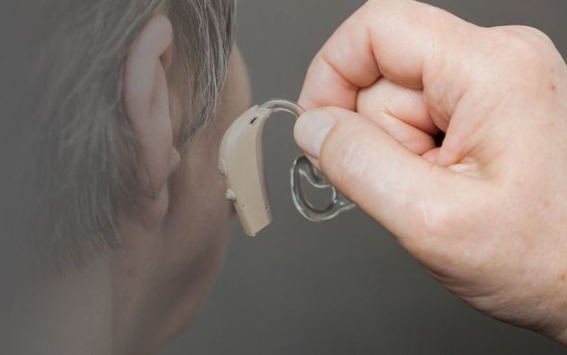 A hand placing a beige behind-the-ear hearing aid onto a person’s ear.