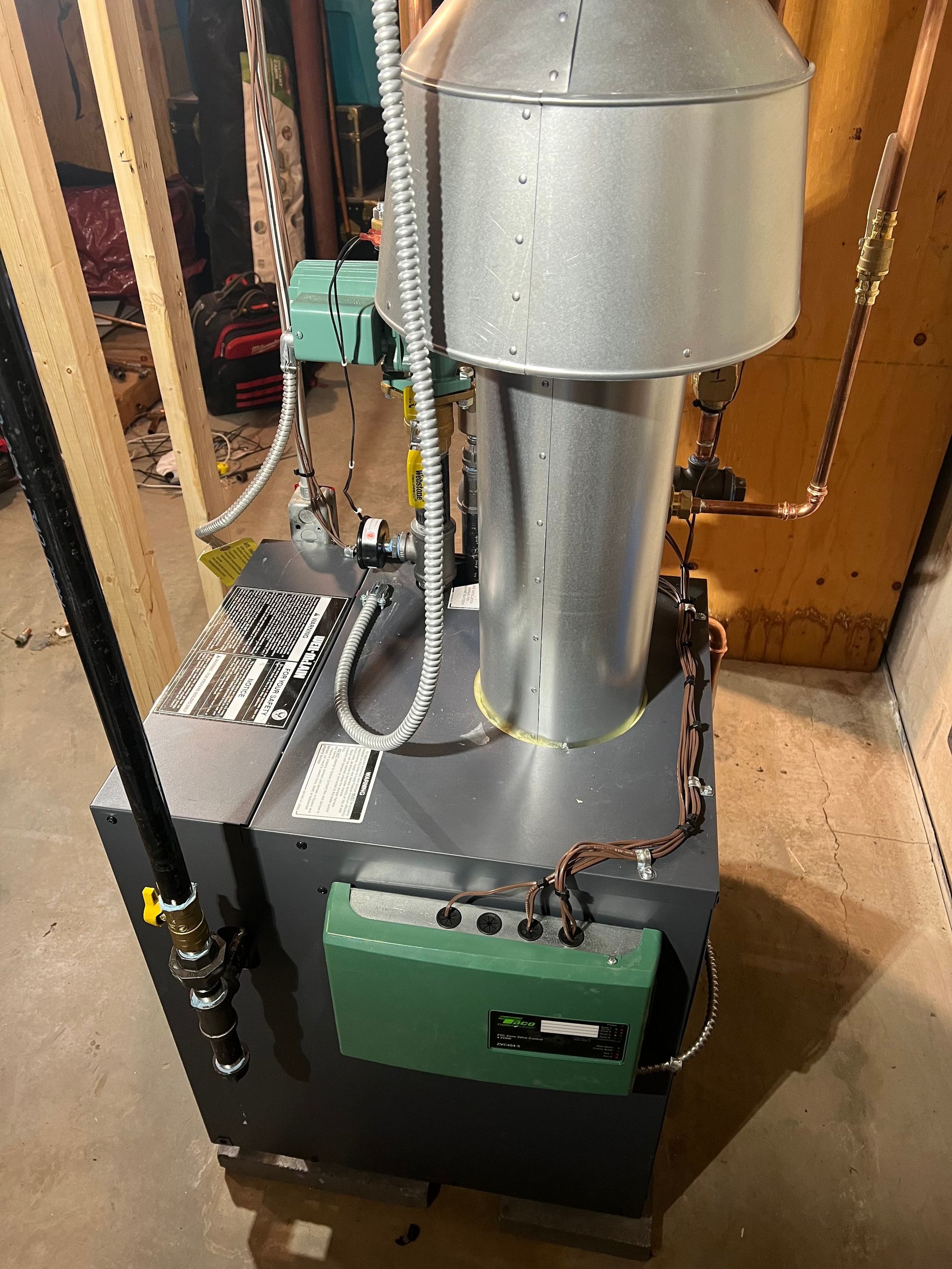 Metal furnace or boiler unit with silver vent pipe and green control box in a workshop corner