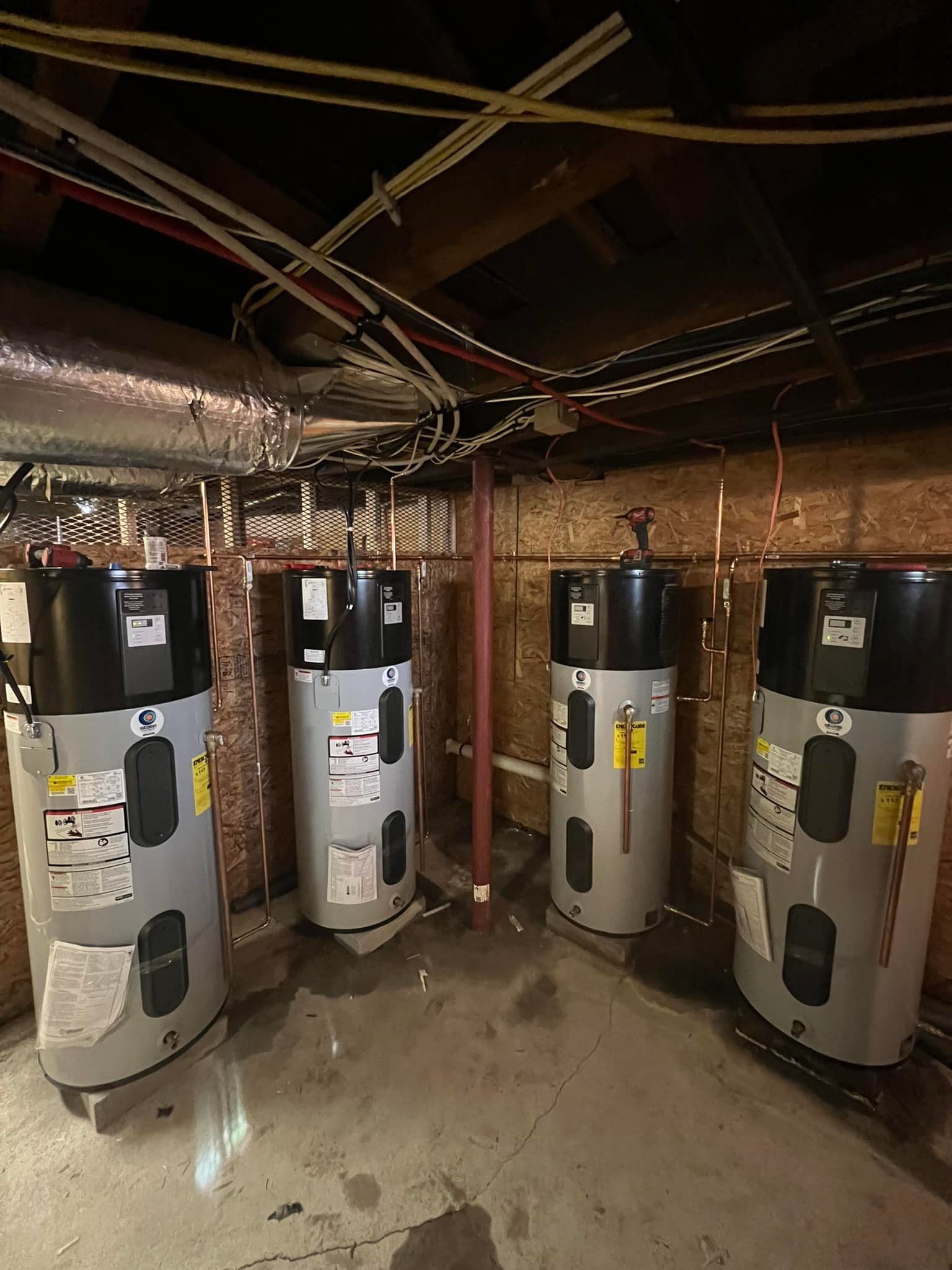 Four silver water heaters in a basement utility room with exposed pipes and brick walls