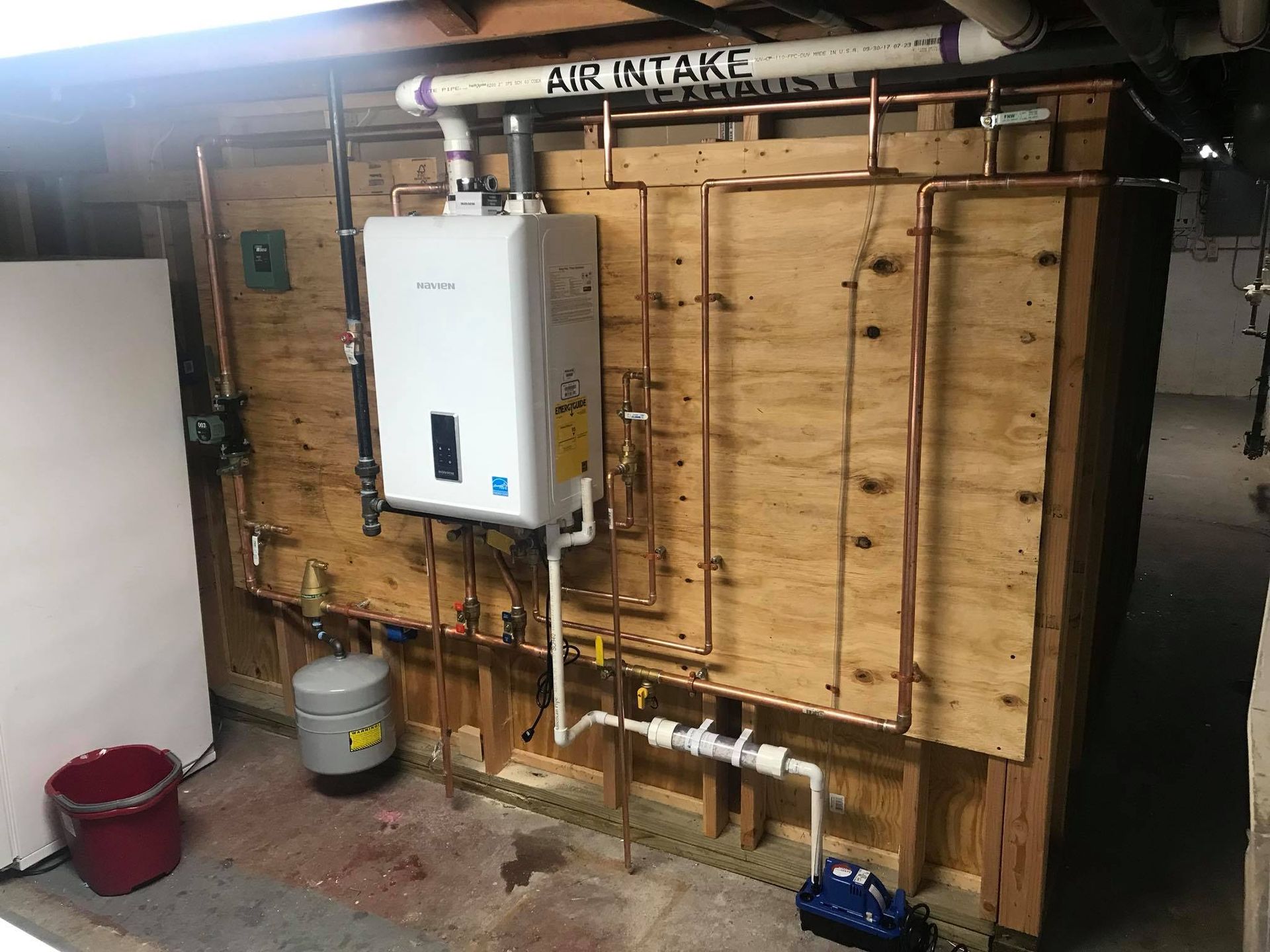 Utility room with a mounted water heater, exposed plumbing, and an air intake sign on a wooden wall.