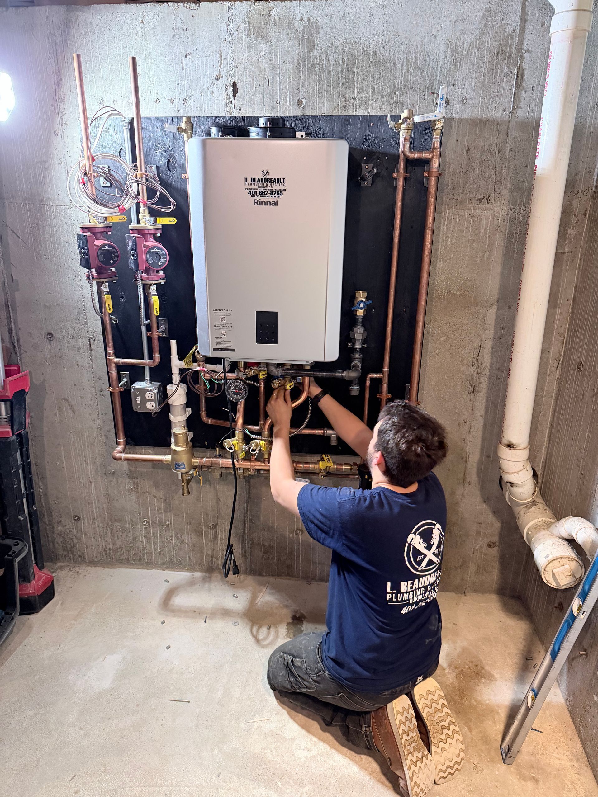 Technician kneeling beside a wall-mounted boiler, working on exposed pipes in an unfinished utility room.