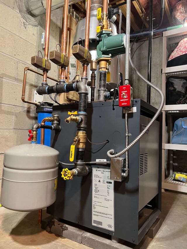 Industrial boiler and piping system in a basement, with gray tank, black unit, and red safety tag.