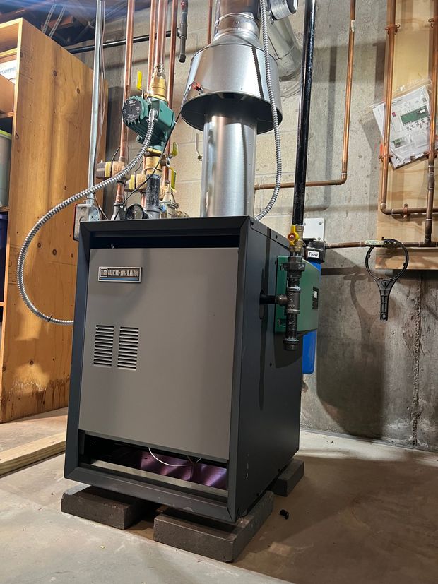 Large gray boiler/furnace in a basement, with metal vent piping and exposed pipes around it.
