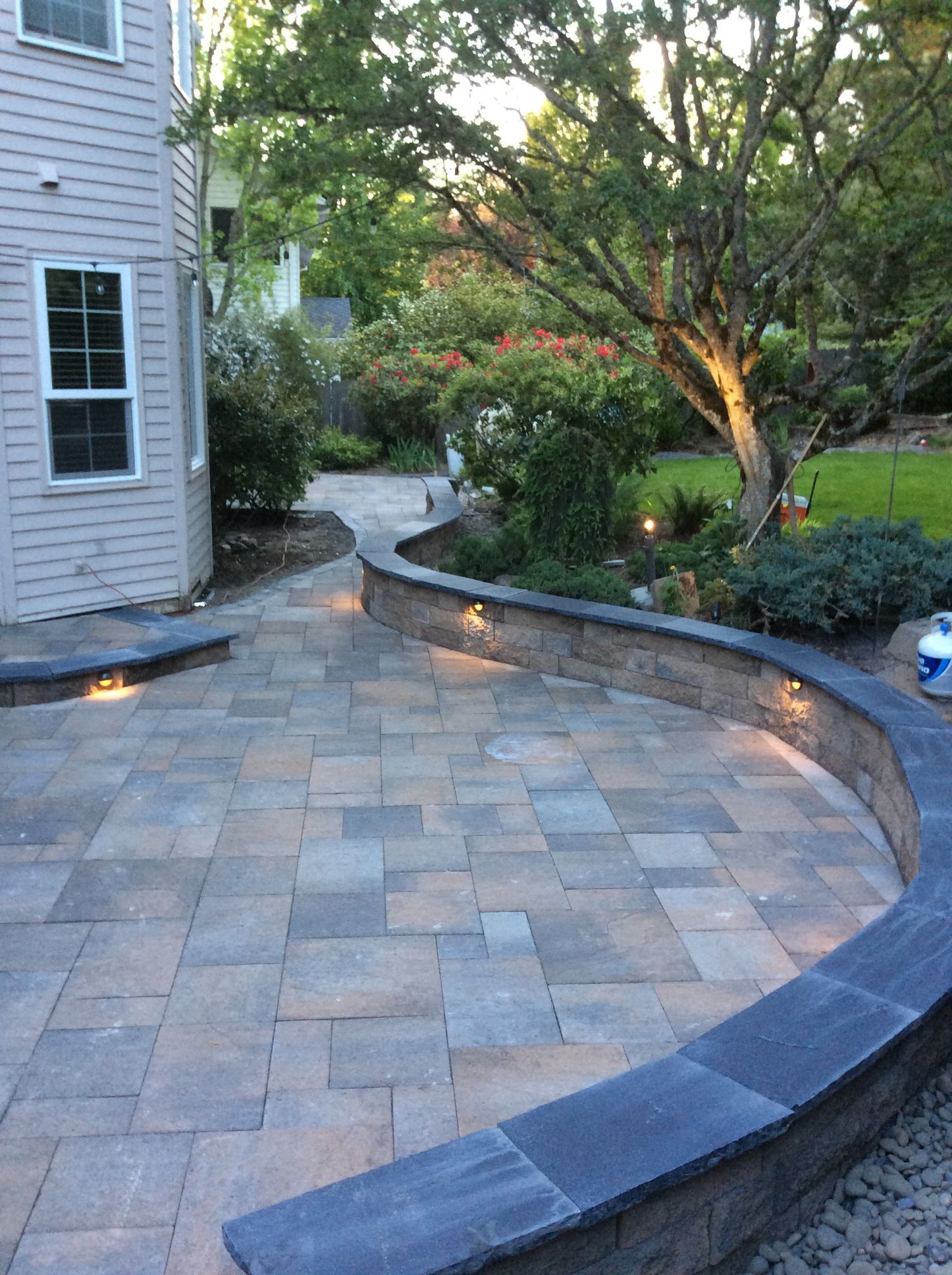 Stone patio with curved retaining wall, landscaped garden beds, and house exterior at dusk