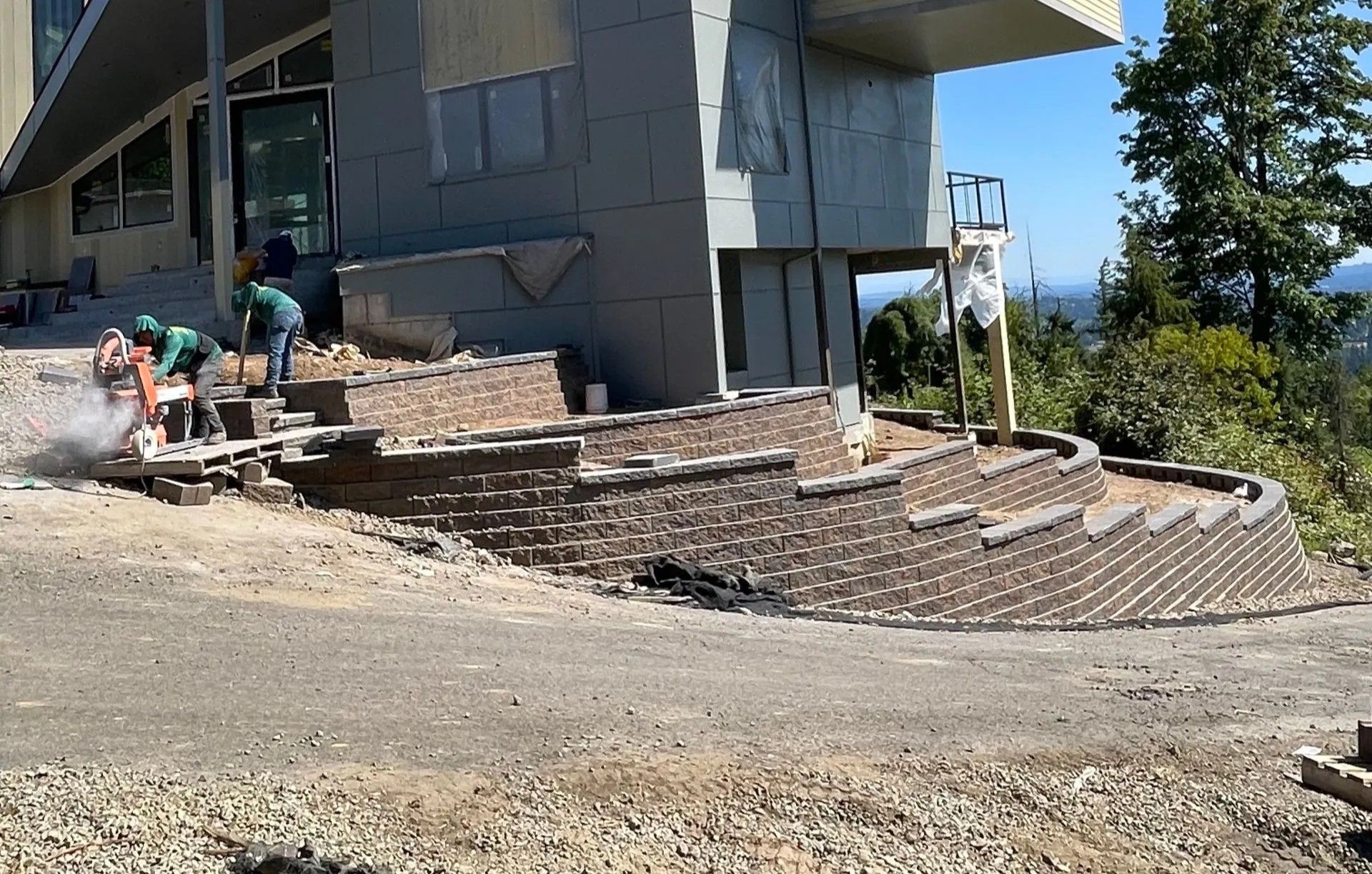 Workers building a stone retaining wall beside a modern building on a dirt slope