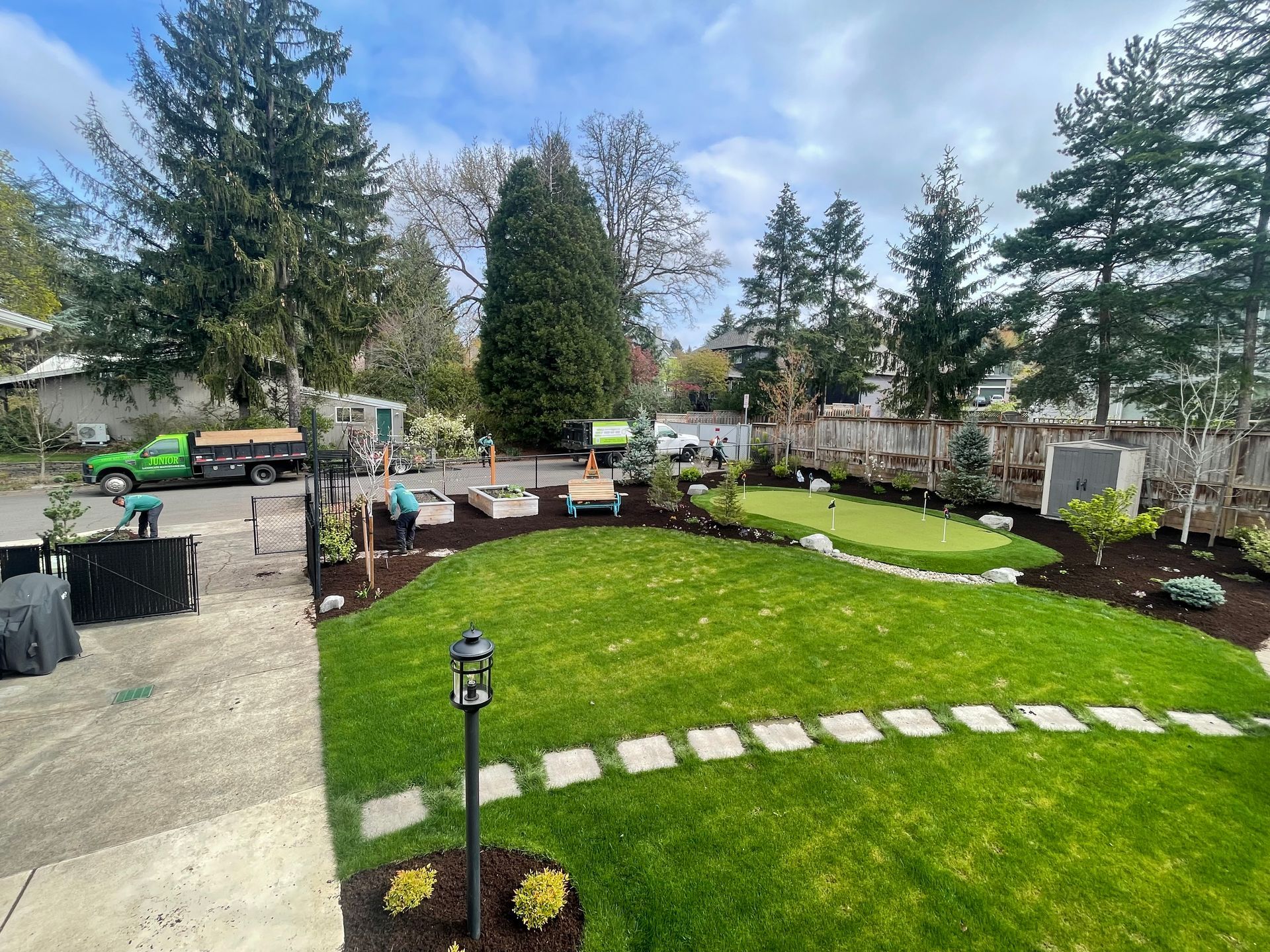 Backyard with green lawn, stone path, patio seating, and tall trees under a cloudy sky