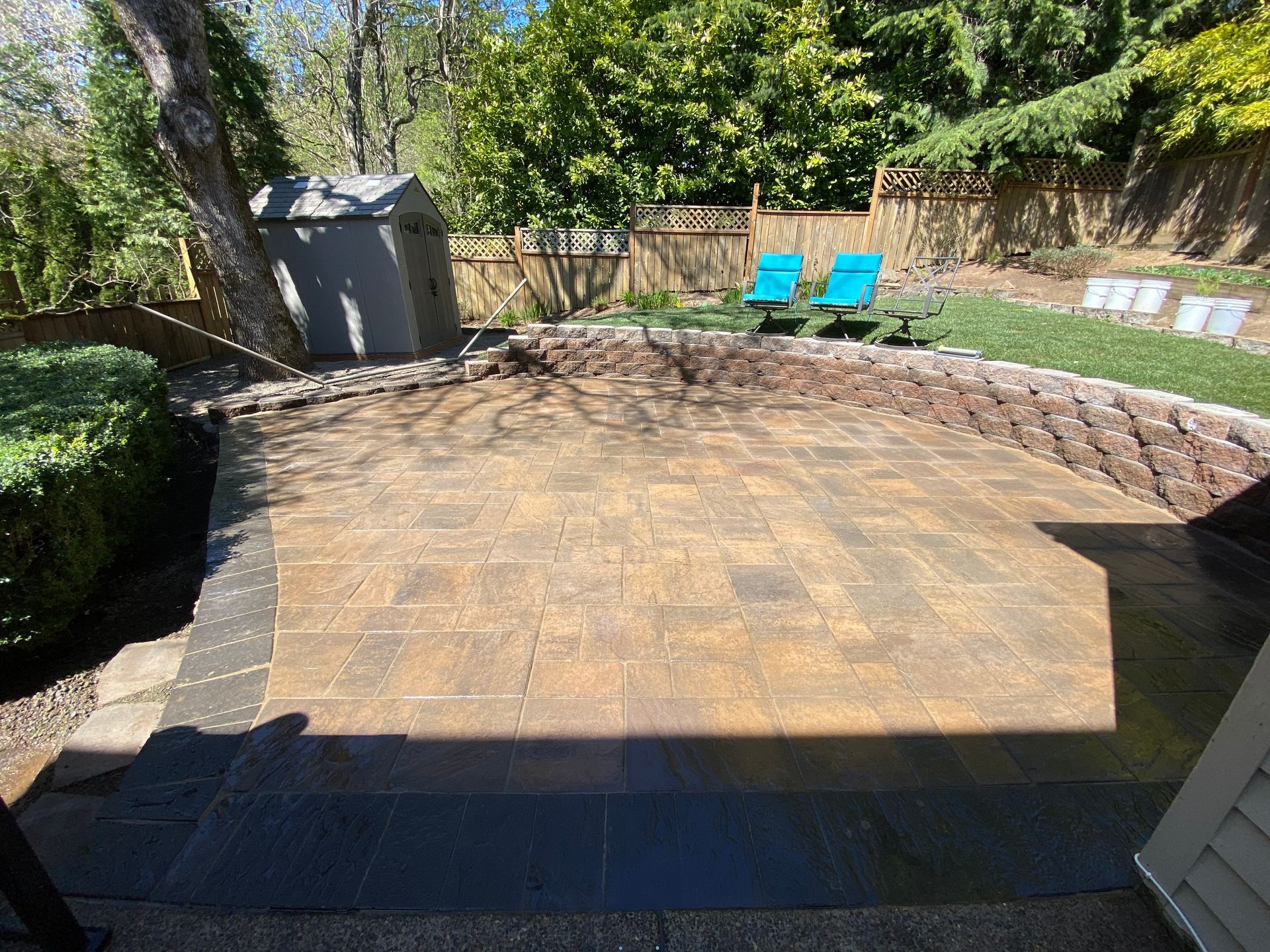 Backyard patio with stone pavers, low retaining wall, blue chairs, and a shed in the shade