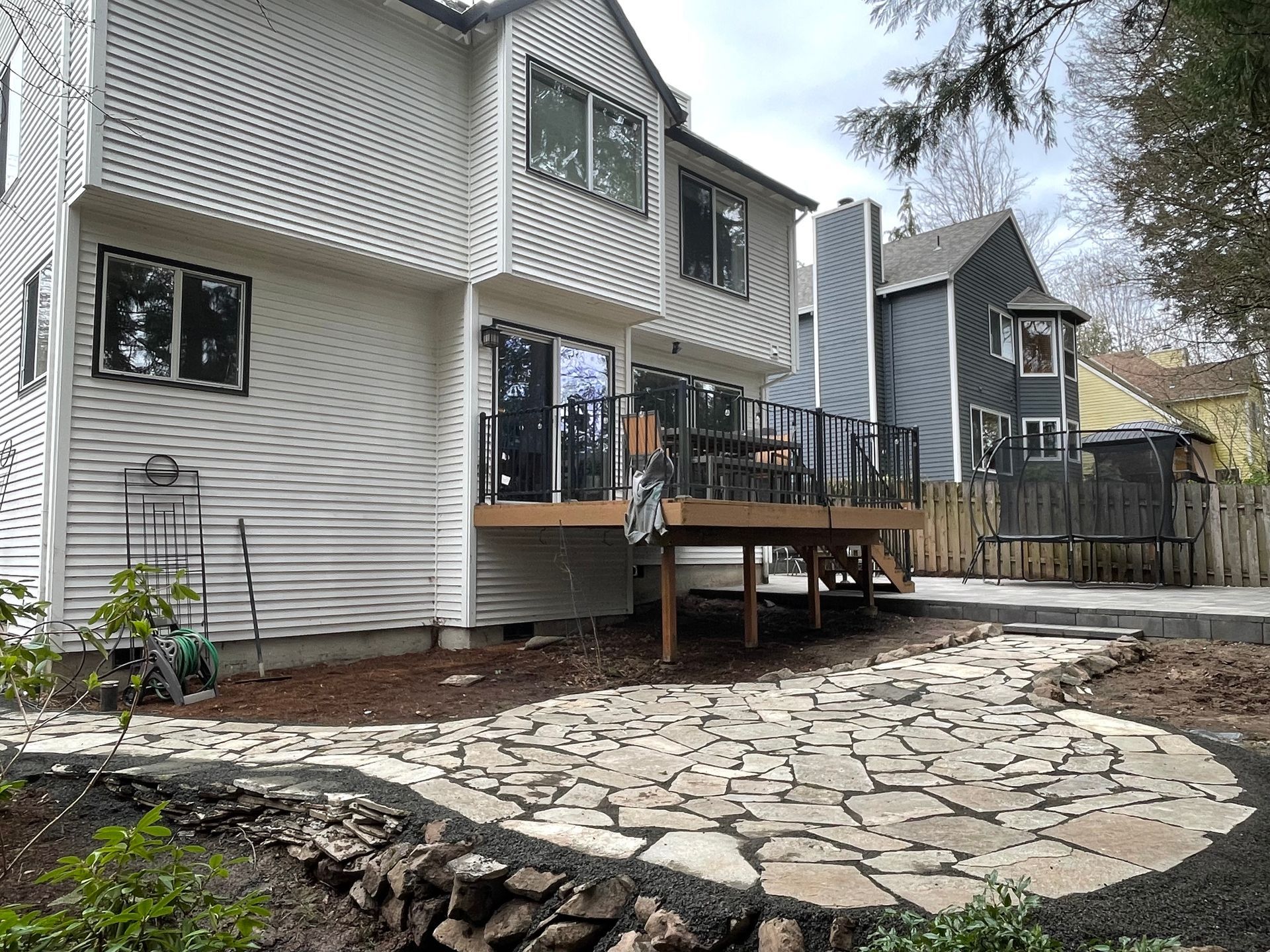 Backyard deck with stone patio and white house siding, surrounded by trees and neighboring homes