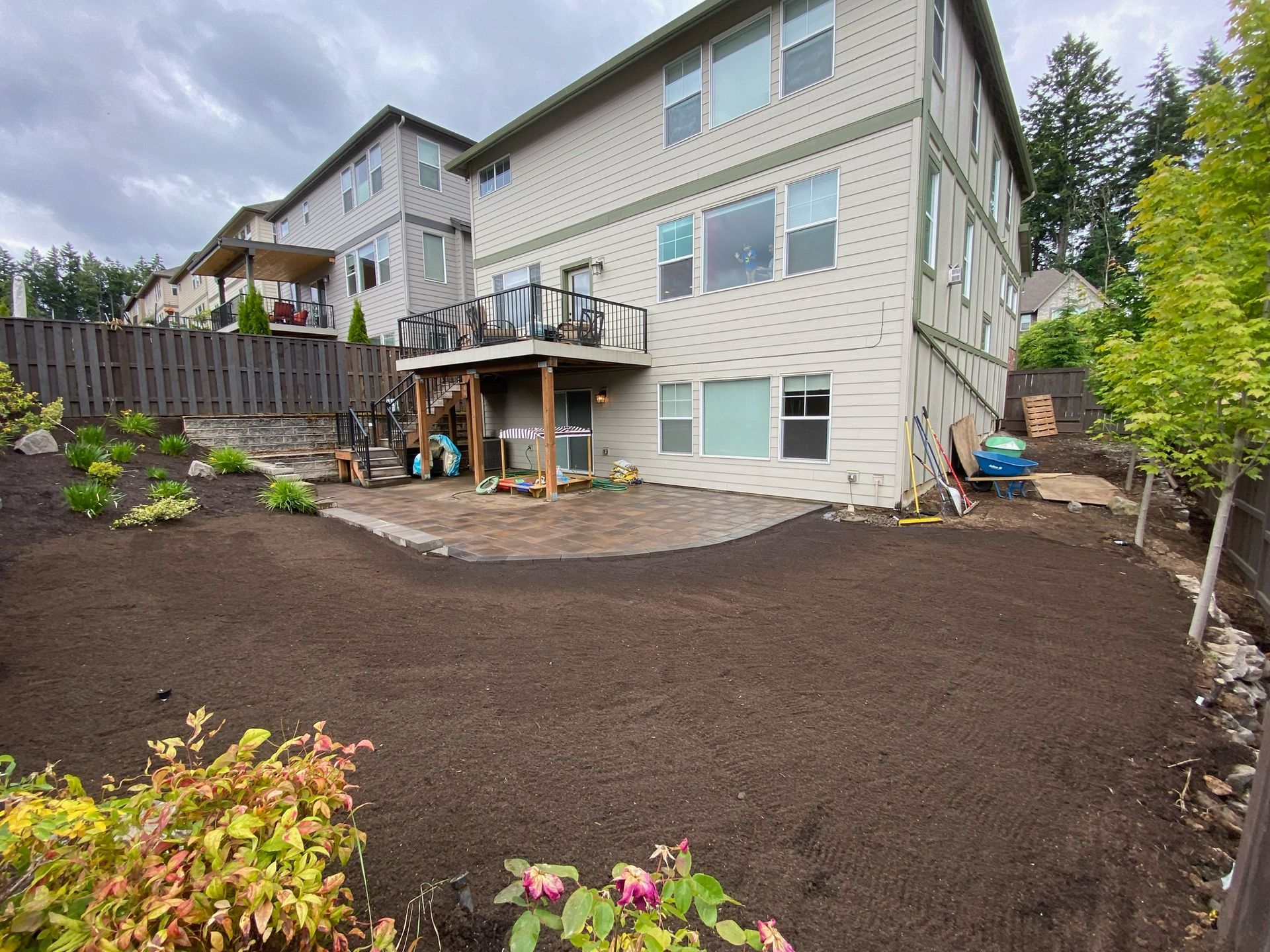 Backyard with fresh dark soil landscaping beside a beige three-story house and wooden deck