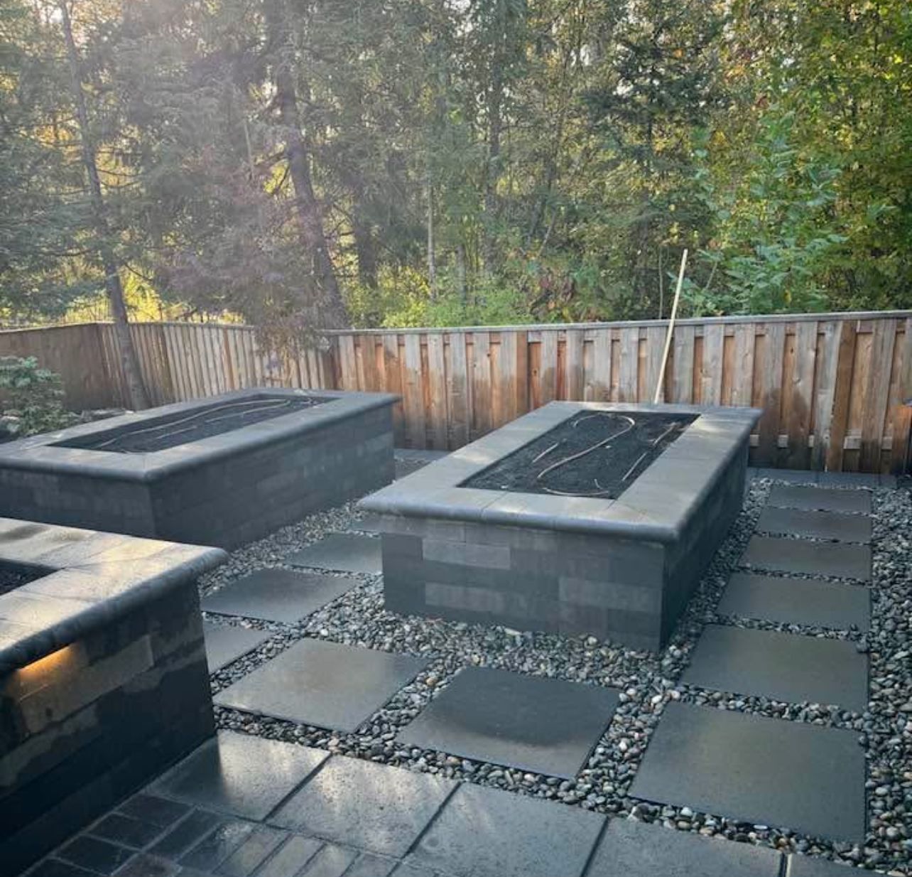 Outdoor patio with two square black fire pits on dark pavers, bordered by a wooden fence and trees.