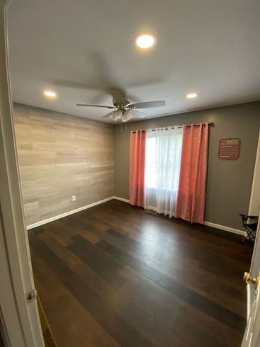 Empty bedroom with dark wood floor, wood-look wall, gray walls, pink curtains, and ceiling fan.